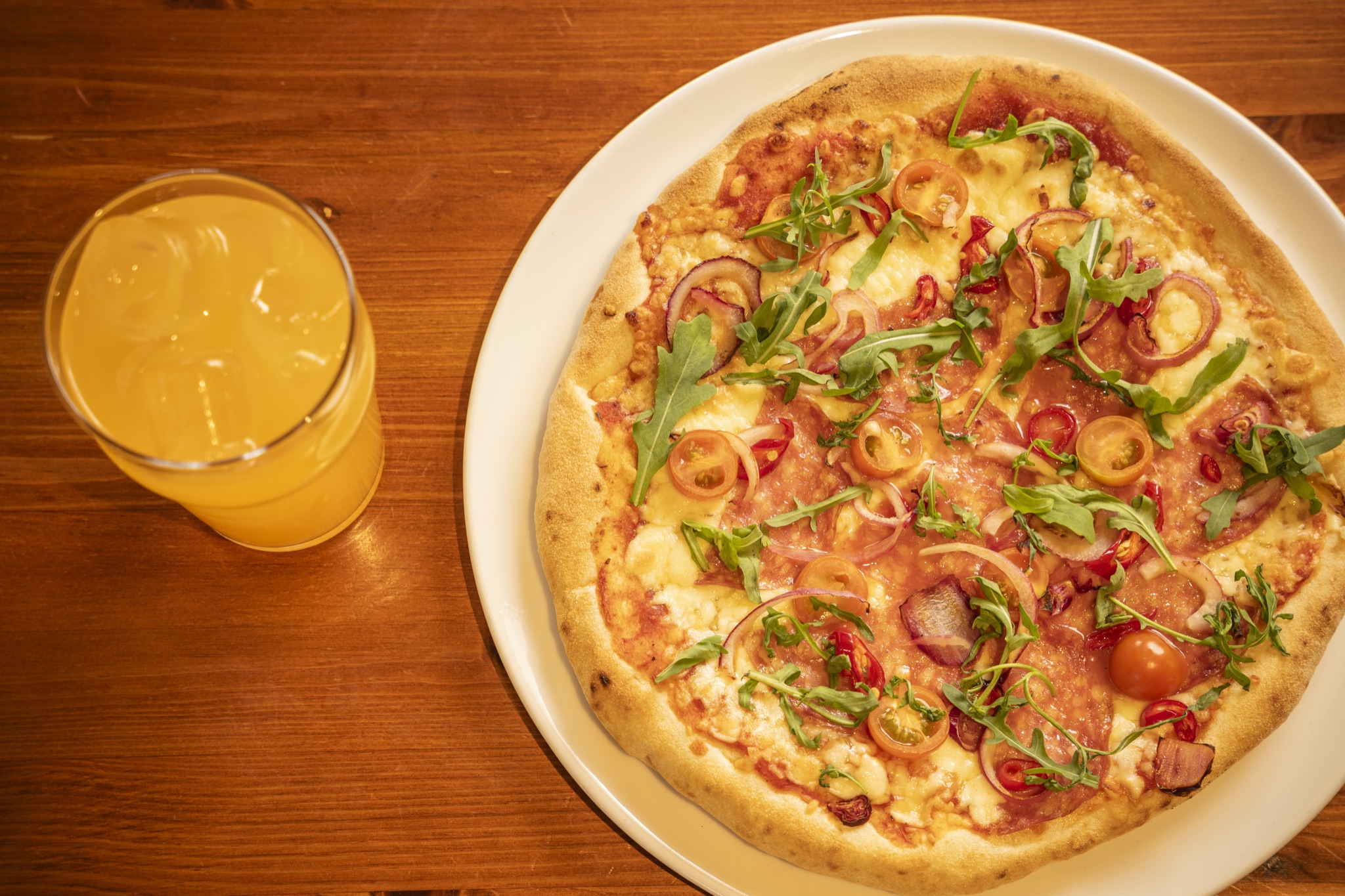 Pizza valkoisella lautasella puupöydällä, vieressä lasi vaaleaa juomaa.