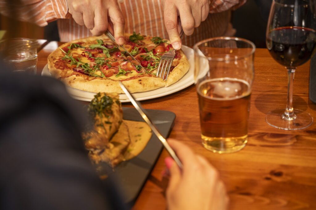 Kaksi ruokailijaa leikkaamassa pizzaa, pöydällä olutlasi ja punaviinilasi.
