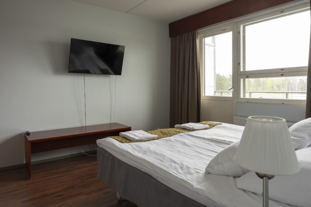 Hotellihuone, jossa leveä vuode, seinätelevisio ja ikkuna puiden reunustamalle pihalle.
