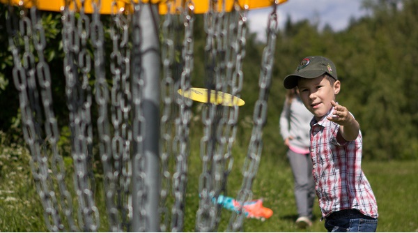 Poika heittämässä frisbeegolfia. Etualalla frisbeegolfkori.